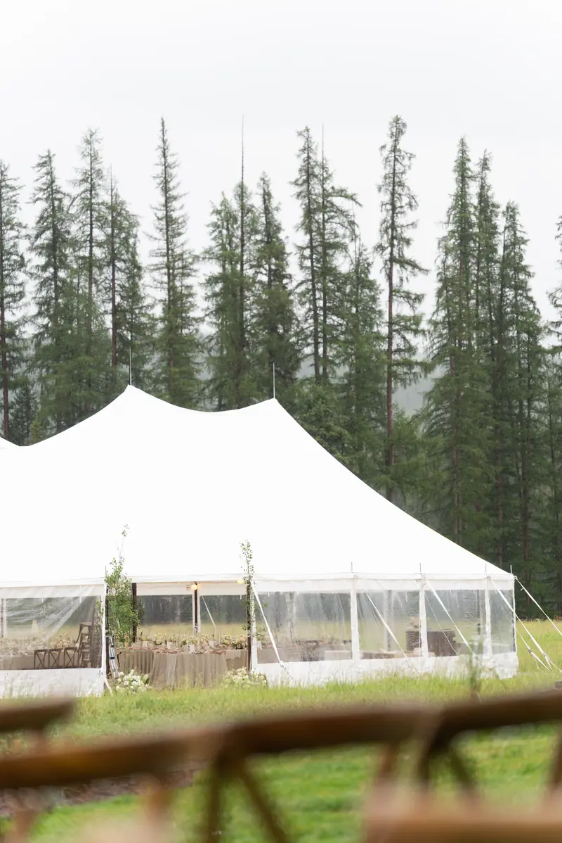 A large white event tent with clear sidewalls in a grassy field against a backdrop of tall pine trees A large white event tent with clear sidewalls in a grassy field against a backdrop of tall pine trees