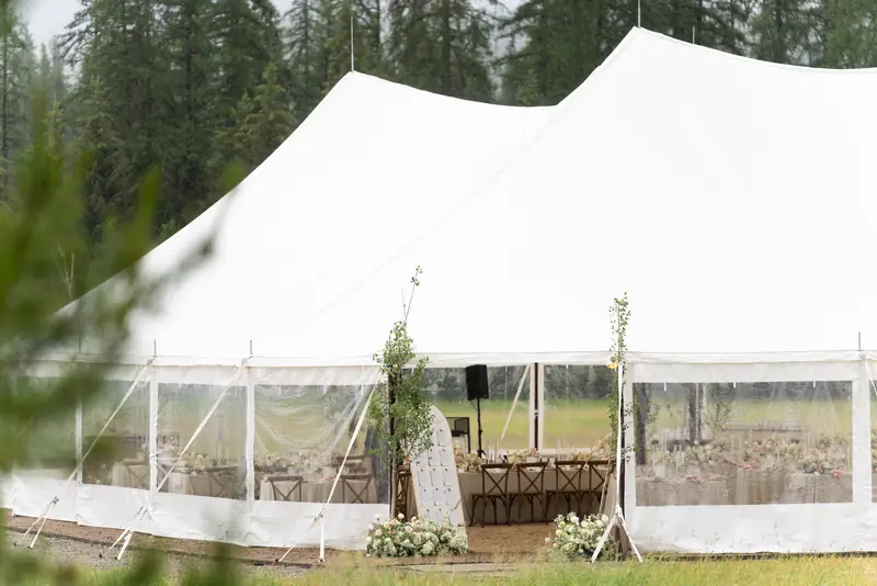 A large white event tent with clear sidewalls set up in a grassy field surrounded by tall trees
