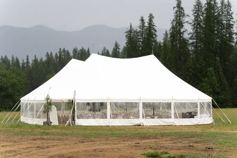 A large white event tent with clear side walls in a field surrounded by tall evergreen trees and distant mountains