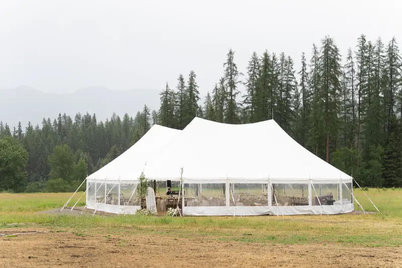 A large white tent with clear walls standing in a field near tall evergreen trees