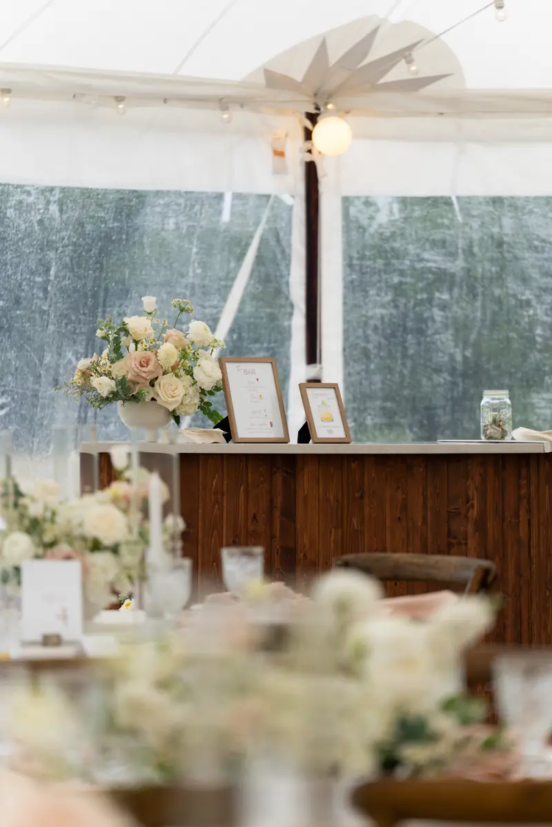 A wood bar and decorated dinner tables inside a white event tent, shot in portrait
