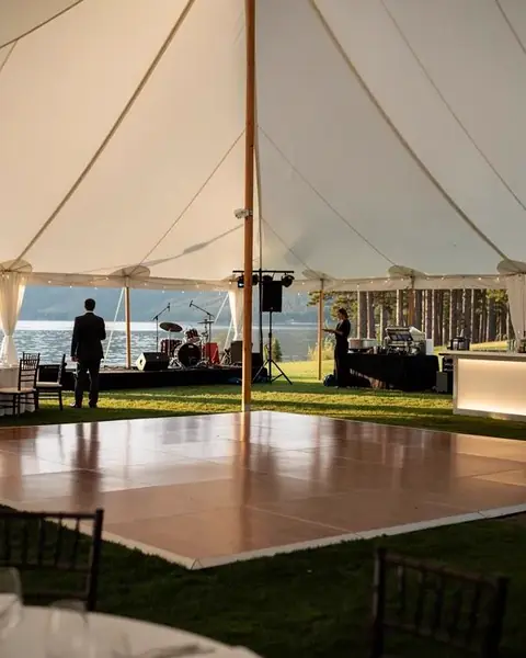 A wood dance floor set in the middle of a white tent by a lake, with a band stage, a lit bar and round tables around it