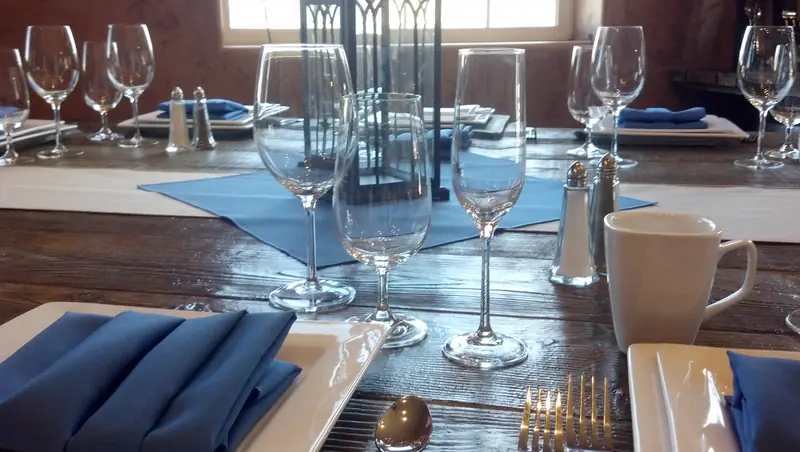 A wood dining table set with wine glasses, white plates and blue napkins, seen low across the top