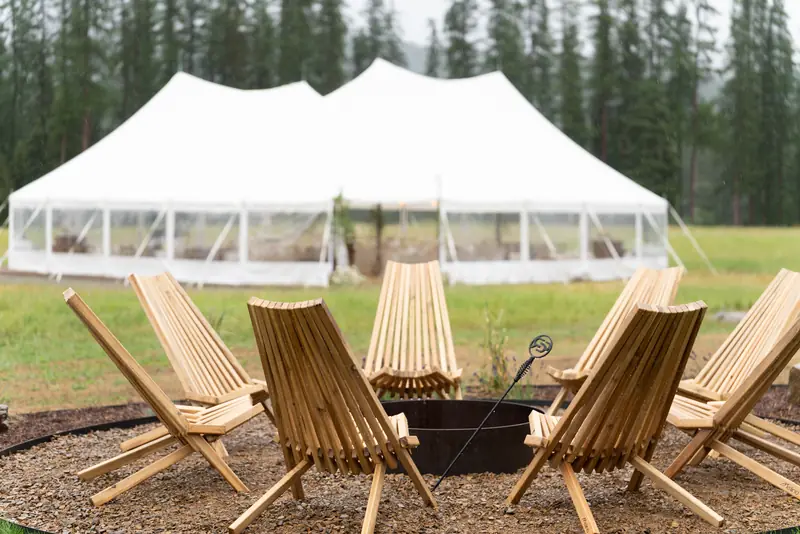 Wooden slatted chairs circle a fire pit on gravel, with a large white event tent in the background Wooden slatted chairs circle a fire pit on gravel, with a large white event tent in the background