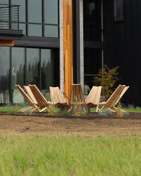 A ring of wooden lounge chairs on a lawn in front of a modern black and timber house